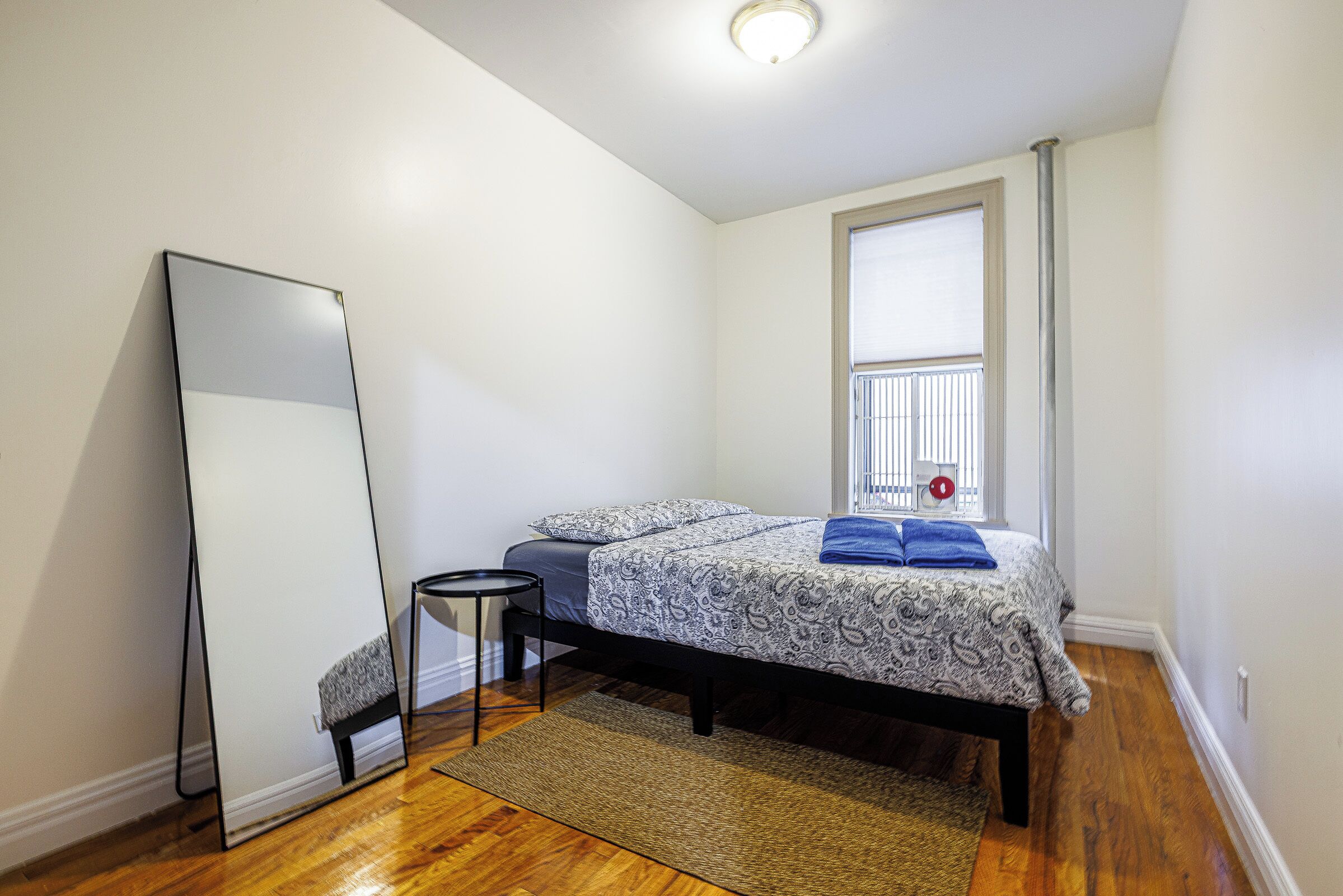 Un dormitorio con cama, espejo de pie, una mesita y una ventana con persianas.