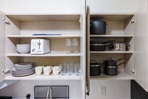 Une cuisine avec des meubles blancs, un grille-pain, des verres à vin, des assiettes, des tasses et des casseroles.