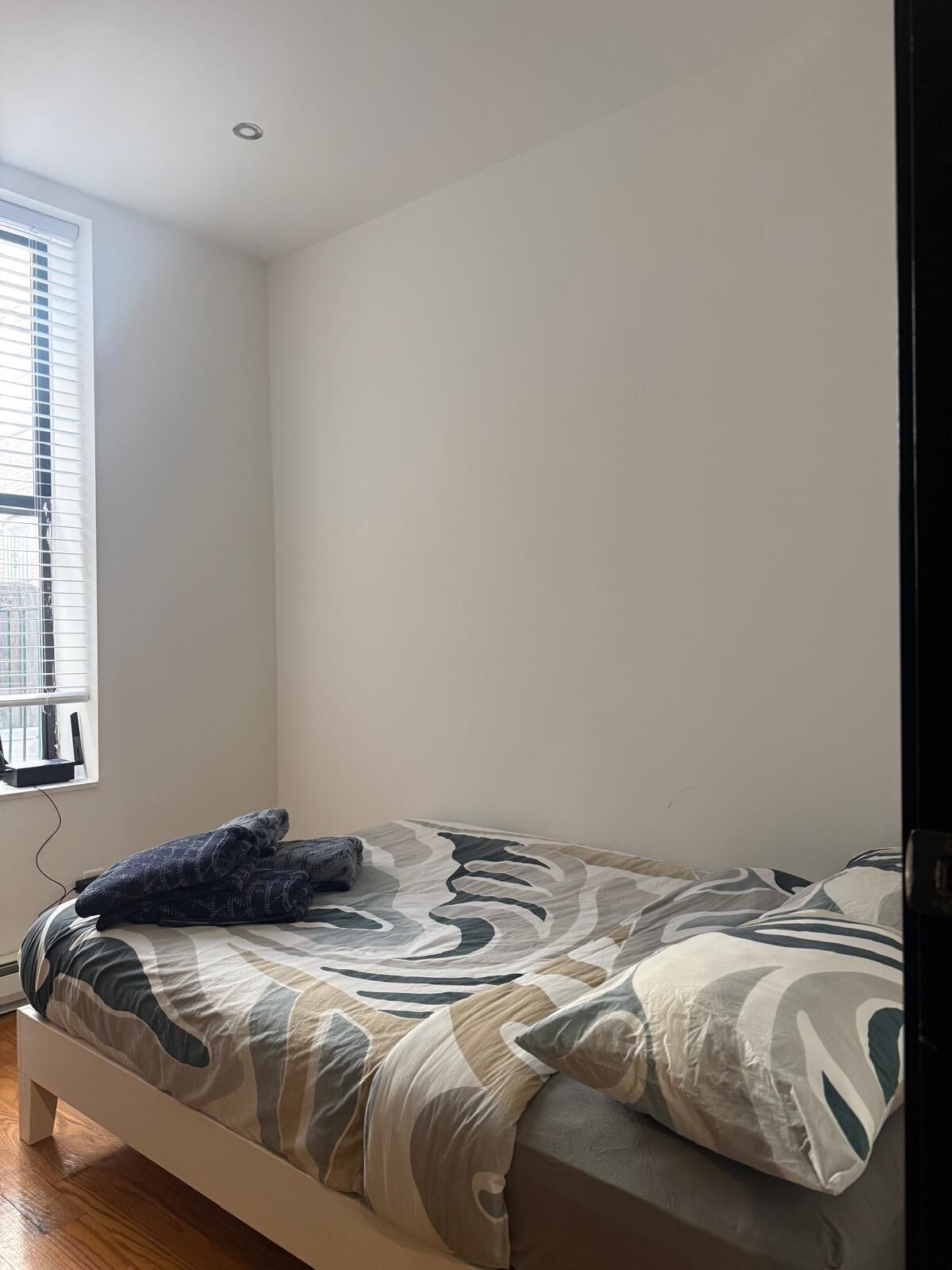 Um quarto com uma cama, uma janela com persianas e uma parede branca.