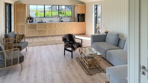 Ein großzügiger Wohnbereich mit grauem Sofa, Korbstühlen und einem Glastisch. Ein Hund ist im Raum, der große Fenster mit Blick ins Freie hat.
