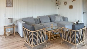 Ein Wohnzimmer mit einem grauen Ecksofa, zwei Korbstühlen, einem kleinen Beistelltisch und einer wandmontierten Klimaanlage.