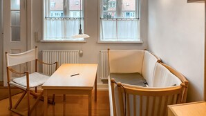Een kamer met een houten vloer, een witte tafel, twee stoelen, een radiator en twee ramen met witte gordijnen.