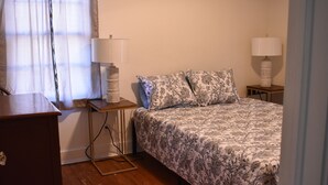 Quarto com cama, duas luminárias de cabeceira, mesa de cabeceira, cômoda de madeira e janela com cortinas finas.