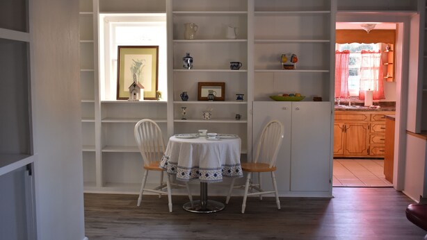 Uma área de refeições com uma mesa redonda e duas cadeiras, um armário branco com prateleiras, e uma cozinha visível através de uma porta aberta.