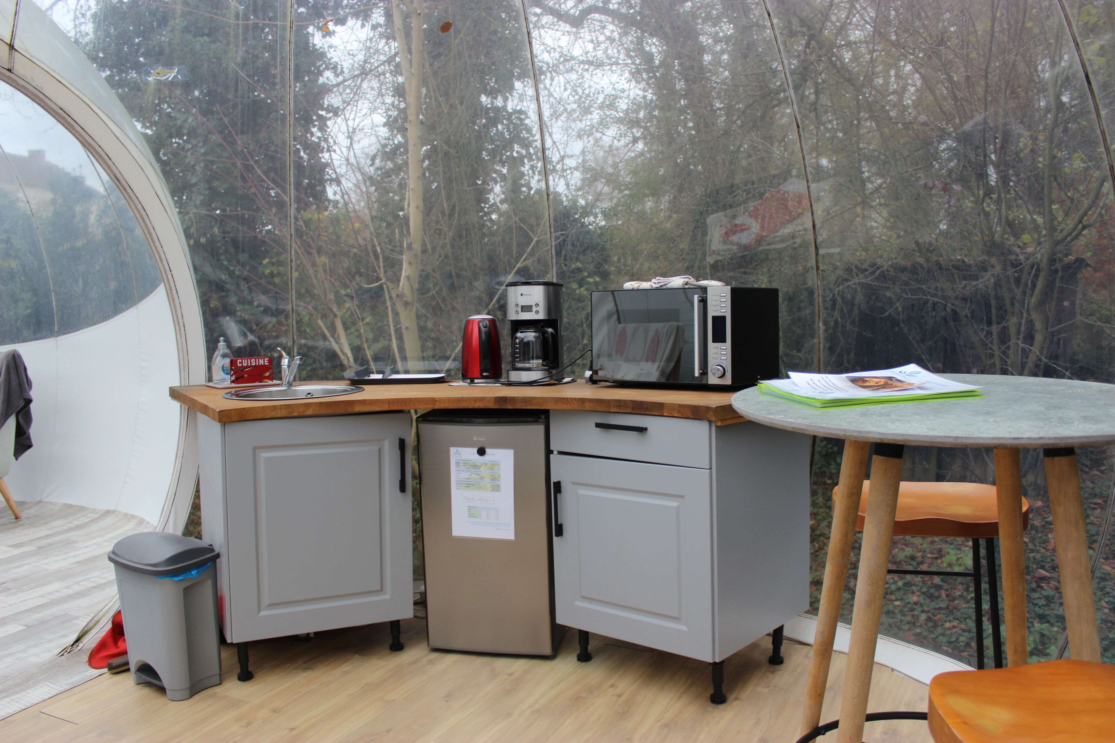 Eine kompakte Küche mit Mikrowelle, Kaffeemaschine und kleinem Kühlschrank. Es gibt eine Holz Arbeitsplatte und einen kleinen Tisch mit einem Stuhl.