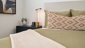 En pænt redt seng med mønstrede puder, en senget lampe og en lille plante på et natbord.