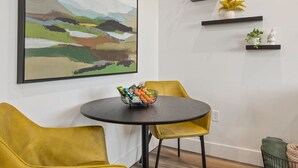 Et moderne spiseområde med et rundt bord, to gule stole, et indrammet abstrakt maleri og minimalistiske hylder med pynt.