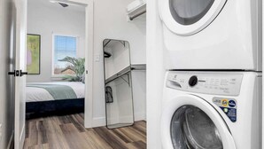 Una lavandería con dos lavadoras de carga frontal apiladas, piso de madera, una cama y una ventana con persianas.