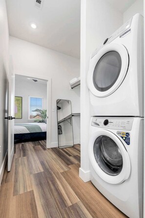 Ein Waschraum mit zwei übereinander gestapelten, frontgeladenen Waschmaschinen, Holzboden, einem Bett und einem Fenster mit Jalousien.