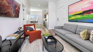 Una sala de estar moderna con un sofá gris, un sillón rojo y una mesa de centro. Hay un cuadro en la pared y una cocina al fondo.