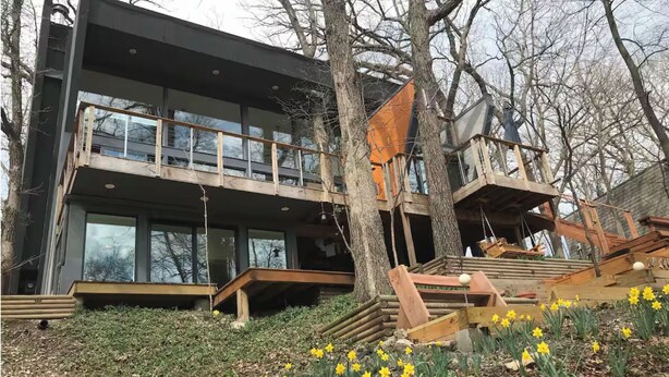 Un bâtiment moderne à deux étages, doté de grandes fenêtres, d’une terrasse en bois et d’un escalier menant à un balcon.