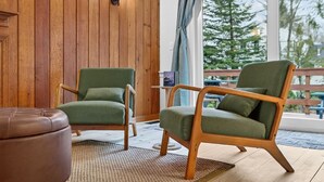 Una habitación con paneles de madera, dos sillones verdes, un reposapiés de cuero marrón y un ventanal con vistas a árboles y un edificio.