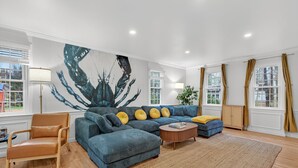 Una sala de estar moderna con un sofá seccional grande, un sillón de cuero y una mesita de centro. En la pared hay un cuadro abstracto de gran tamaño y una ventana con cortinas.