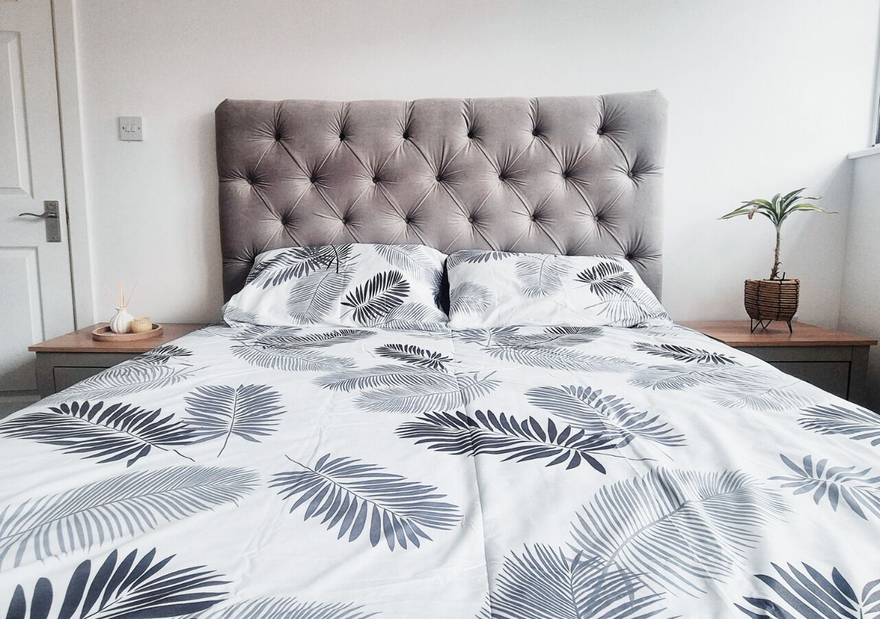 A bedroom with a tufted headboard, a bed with a leaf-patterned duvet, a bedside table with a plant, and a nightstand with a candle.