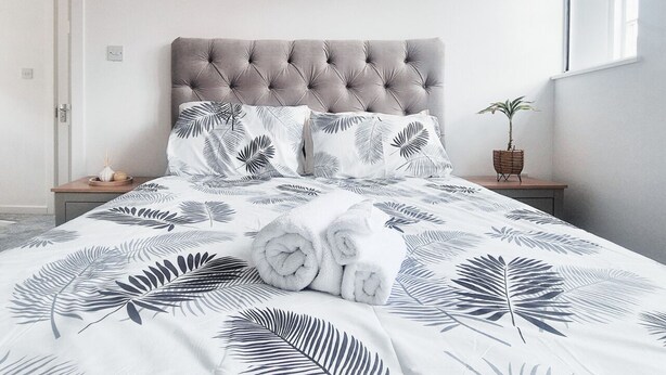 Ein Bett mit einem gepolsterten Kopfteil, einer zudeckenden Bettwäsche mit Blumenmuster und aufgerollten Handtüchern.