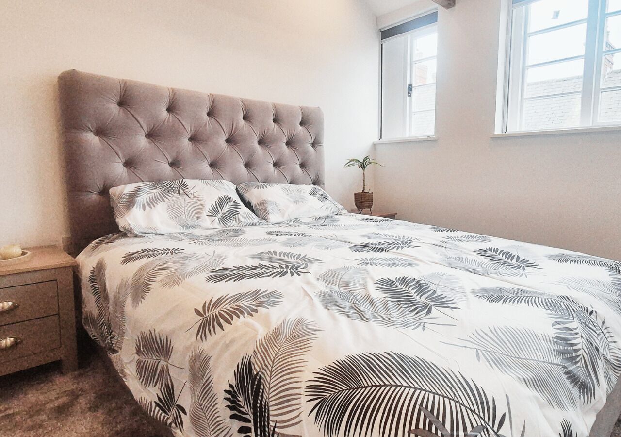 Une chambre à coucher avec une tête de lit capitonnée, un lit recouvert d’une couvre-lit à motifs de feuilles, une table de chevet avec une lampe et une fenêtre avec des stores.