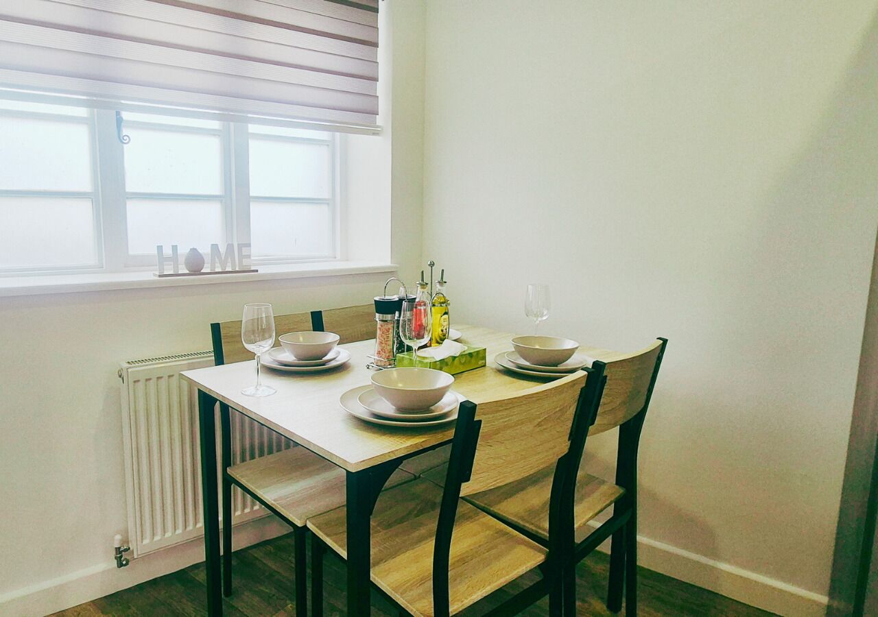 A dining table with four chairs, a window with blinds, a radiator, and a 'HOME' sign on the wall.