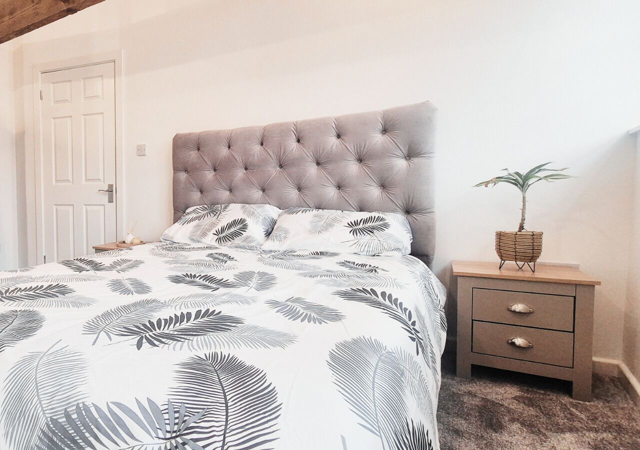 Ein Schlafzimmer mit einer gepolsterten Kopfteile, einem Bett mit Bettwäsche mit Blattmuster, einem Nachttisch mit Schubladen und einer Topfpflanze.