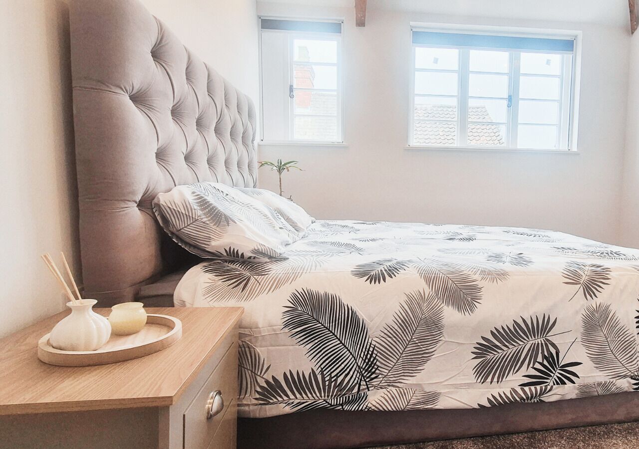 Une chambre à coucher avec une tête de lit capitonnée, un lit recouvert d’une couvre-lit à motifs de feuilles, une table de chevet agrémentée d’une petite plante et une fenêtre donnant sur l’extérieur.