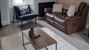 Una sala de estar moderna con un sofá de cuero, un sillón oscuro, una mesa de centro y un radiador. Hay cuadros enmarcados en la pared y una ventana con persianas.