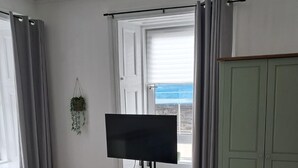 Una habitación con televisión de pantalla plana sobre un soporte tipo trípode, una ventana con cortinas, un armario verde y una planta colgante.