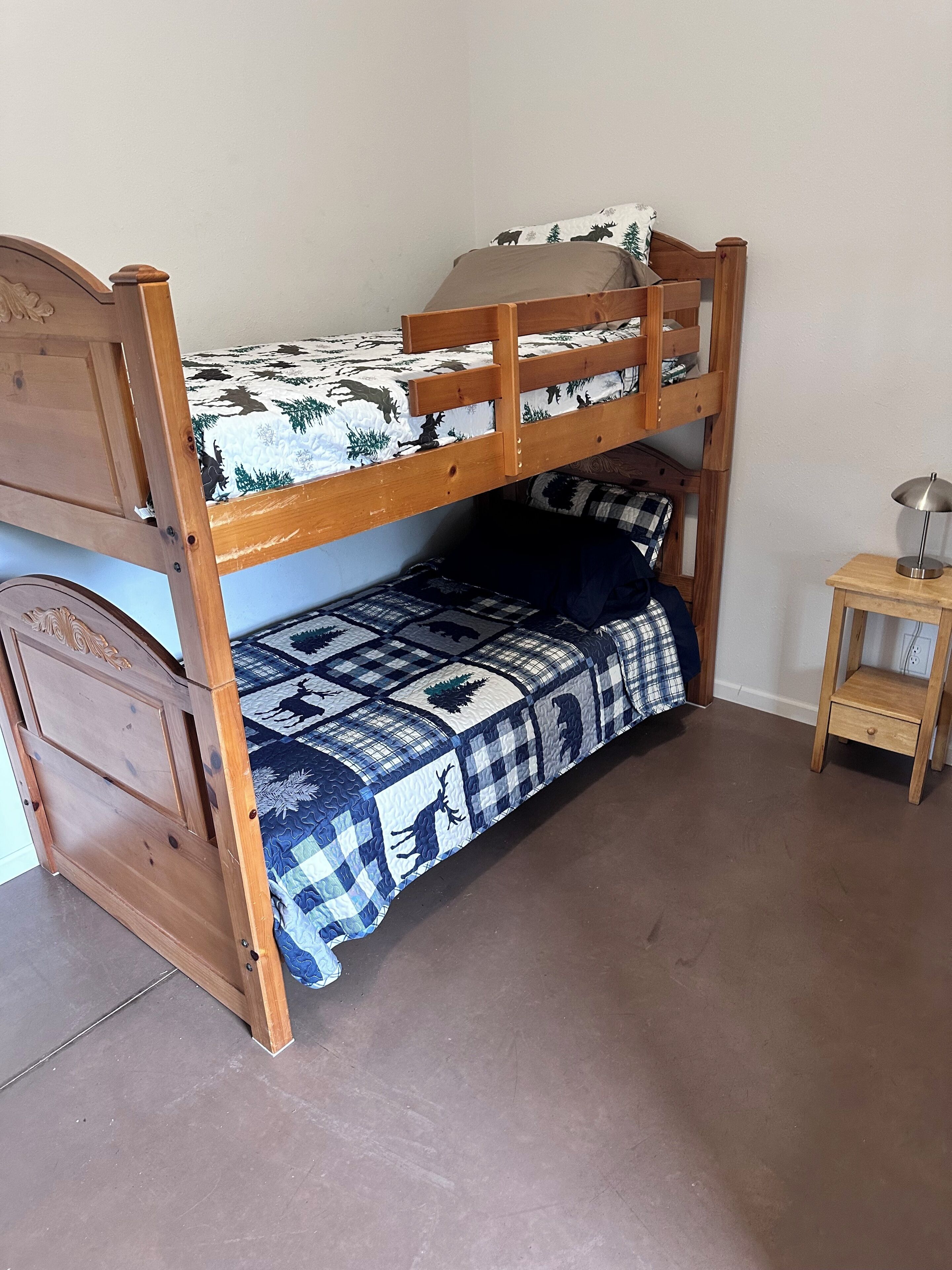 Una cama litera con colcha estampada, una mesita de noche de madera y una pared lisa.