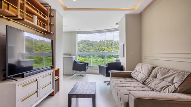 A modern living room with a large TV, a sofa, a coffee table, and a view of a forest through large windows. - Your Complete Stay Just Minutes From the sea in BC Stls (Balneário Camboriú)