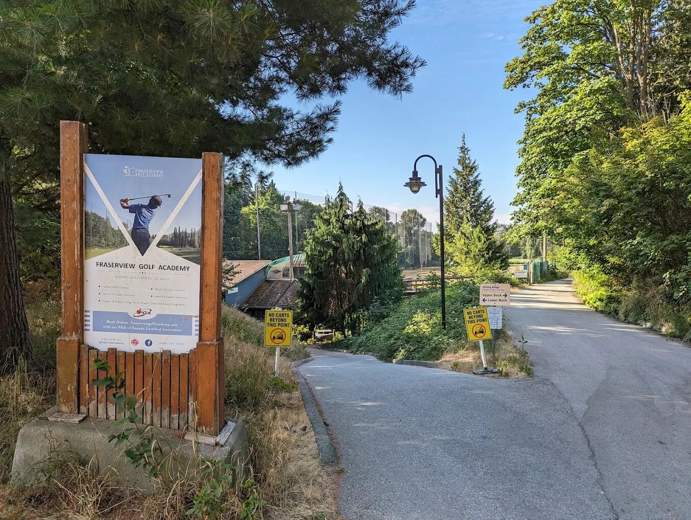 A sign for Fraserview Golf Academy with a wooden fence and a pathway leading to a road.