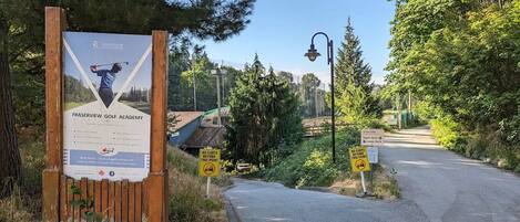 A sign for Fraserview Golf Academy with a wooden fence and a pathway leading to a road.