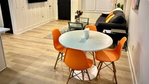 En moderne stue med et rundt glasbord og orange stole, en sort sofa med puder, og et fladskærms-TV monteret på væggen.