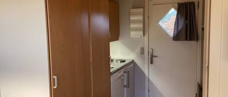 Una habitación compacta con un armario de madera, una pequeña kitchenette y una puerta con ventanal.