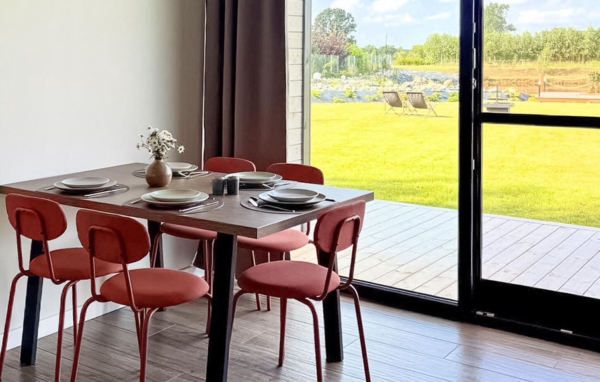 A dining area with a table set for four, red chairs, and a view of a grassy outdoor area through a large window.