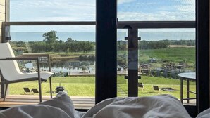 A bedroom with a large window overlooking a lake, a deck with outdoor furniture, and a view of a park and buildings in the distance. - Cozy home in Sianowo with house sea view (Sianowo)