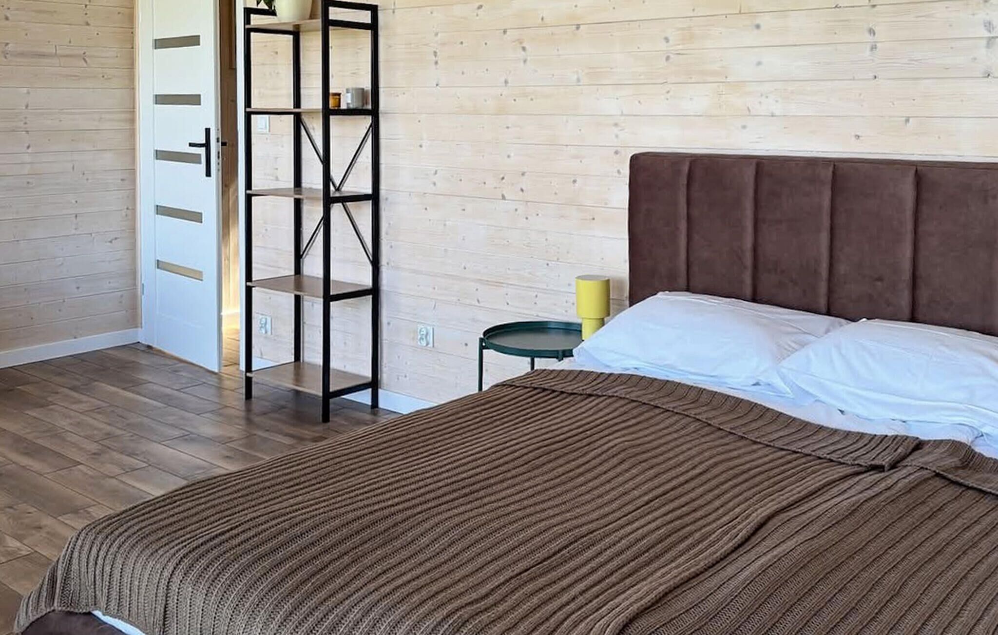 A bedroom with a bed, a wooden wardrobe, a small table with a lamp, and a wooden floor.
