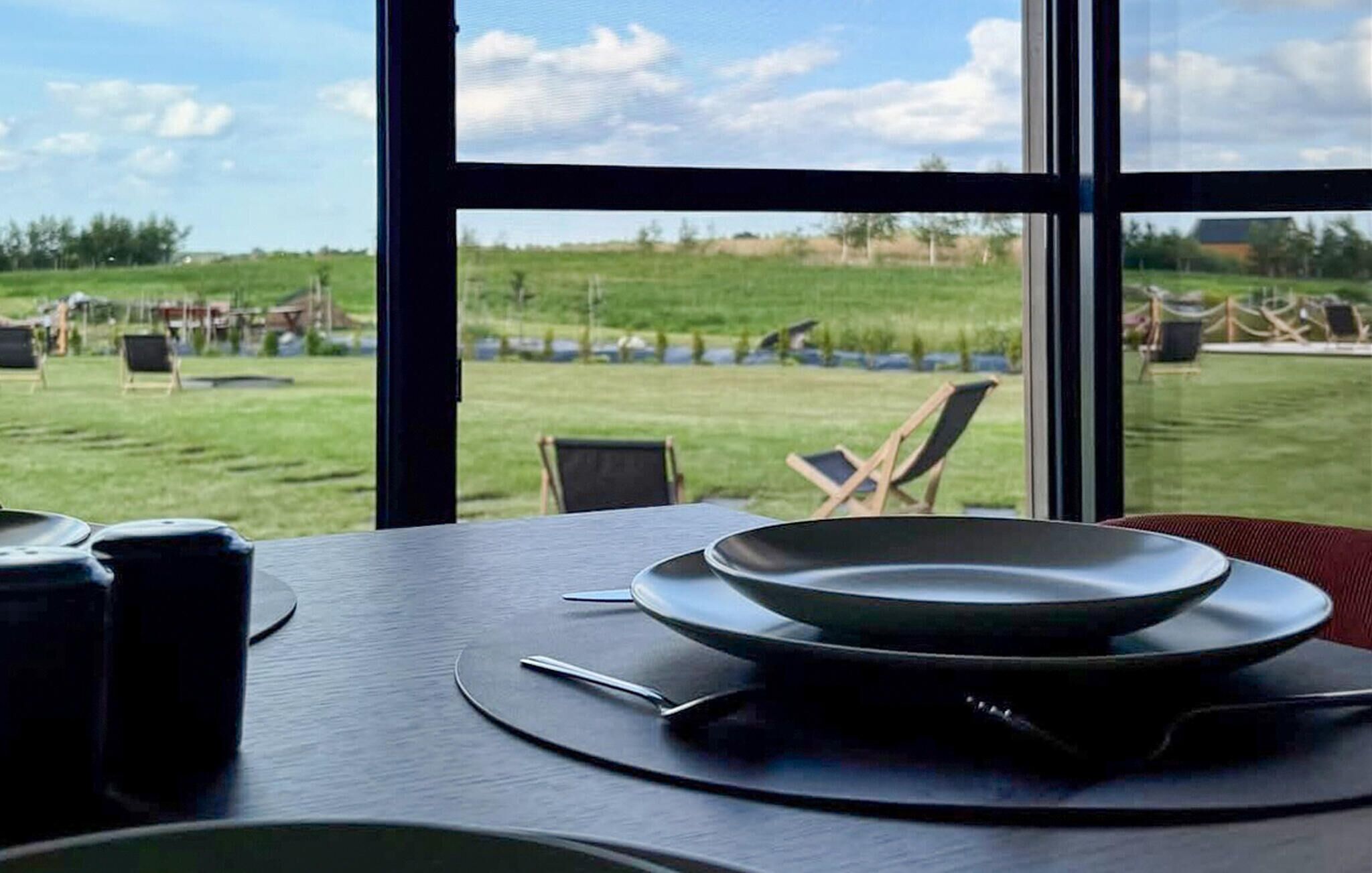 A dining table with plates and cutlery, a window with a view of a grassy field and chairs outside.
