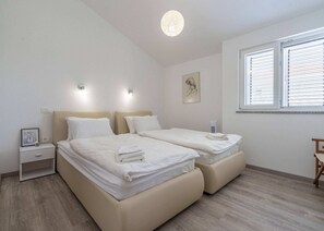 Ein Doppelzimmer mit zwei Betten, Nachttischen, einer Wandleuchte, einem Fenster mit Jalousien und einem gerahmten Bild an der Wand.