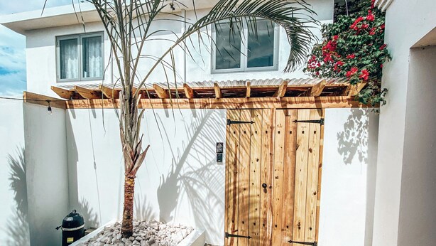 Un bâtiment blanc avec une porte en bois et un petit jardin agrémenté d’un palmier et de petits galets blancs.