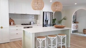 Een moderne keuken met witte kasten, een groot eiland met krukken, een roestvrijstalen koelkast en een hanglamp.
