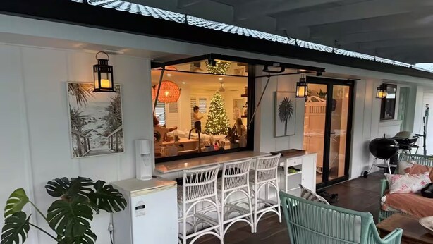Een knus interieur met een witte koelkast, een witte rieten stoel, een blauwgroen ledikant en een kerstboom zichtbaar door een glazen deur.