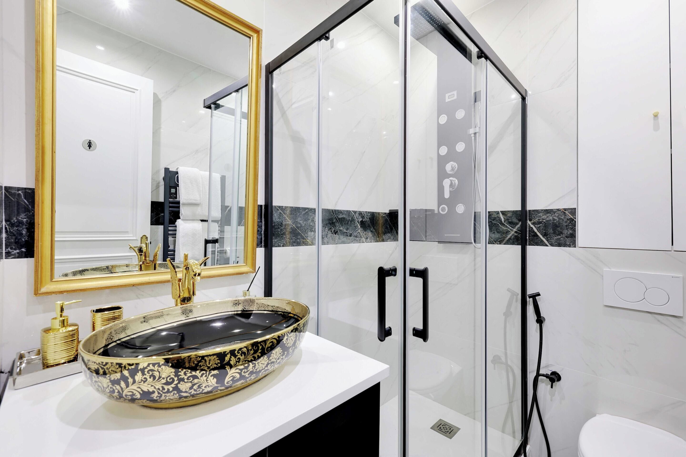 Un baño con ducha envuelta en vidrio, un lavamanos ornamentado en dorado y negro, y un espejo grande con marco dorado.