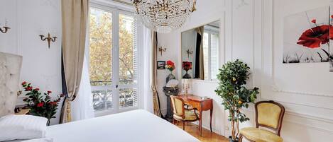A bedroom with a large bed, a chandelier, a chair, a table, and a potted plant.