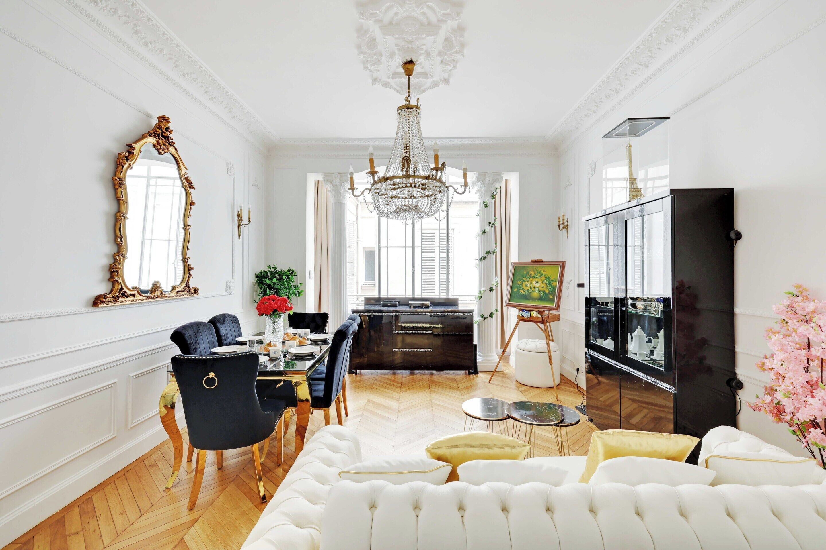 Een ruime kamer met een grote witte bank, een zithoek met een zwarte fauteuil, een kroonluchter en een zwarte kast met glazen deuren.