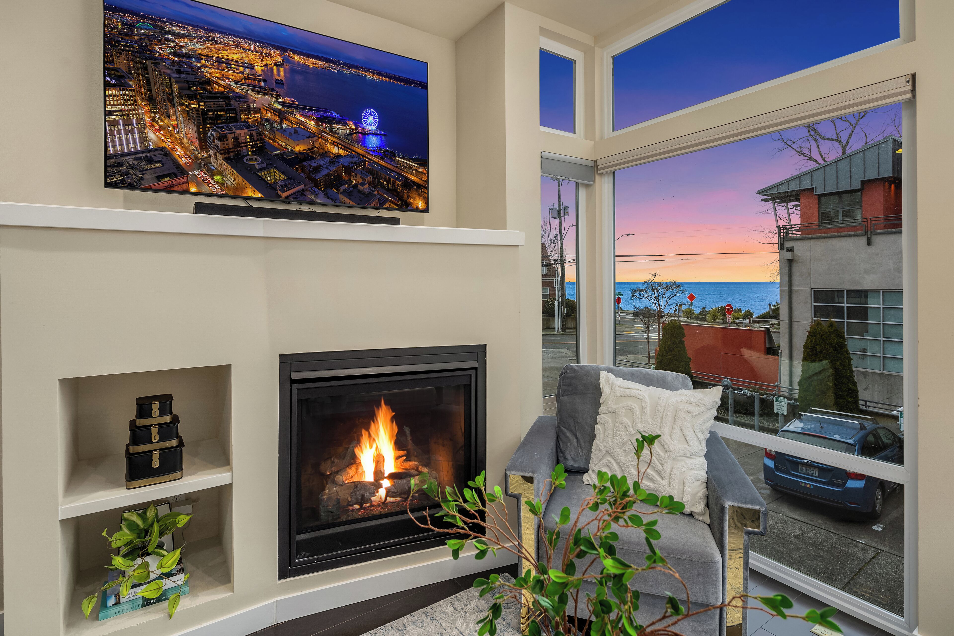 A modern living room with a fireplace, a television mounted on the wall, and a large window offering a view of the cityscape and the ocean.