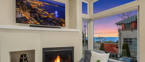 A modern living room with a fireplace, a television mounted on the wall, and a large window offering a view of the cityscape and the ocean.