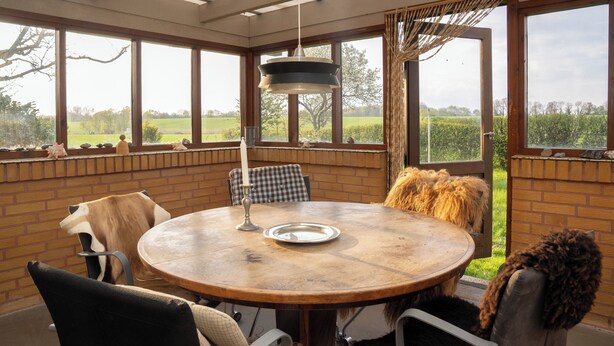 Una mesa redonda de madera con sillas, una lámpara colgante y vistas a un paisaje verde a través de amplios ventanales.