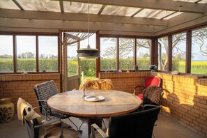 A sunlit room with a round wooden table, chairs, and large windows offering a view of a green field.
