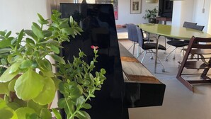 Un interior moderno con un piano negro, plantas verdes, un comedor con mesa y sillas, y un techo de madera.