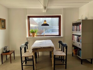 Et værelse med et bord og stole, en bogreol, et vindue med en plante og et billede i ramme på væggen.
