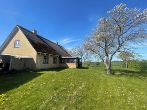 Ein eingeschossiges Haus mit geneigtem Dach, einer kleinen Veranda und einem Baum mit weißen Blüten im Vordergrund.