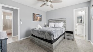 Une chambre à coucher comprenant un lit, un ventilateur de plafond, une table de chevet, une commode, une lampe fixée au mur et une porte donnant sur une salle de bain.